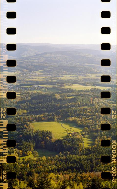 An aerial view shows a vast landscape of rolling hills, forests, and fields under a bright sky.