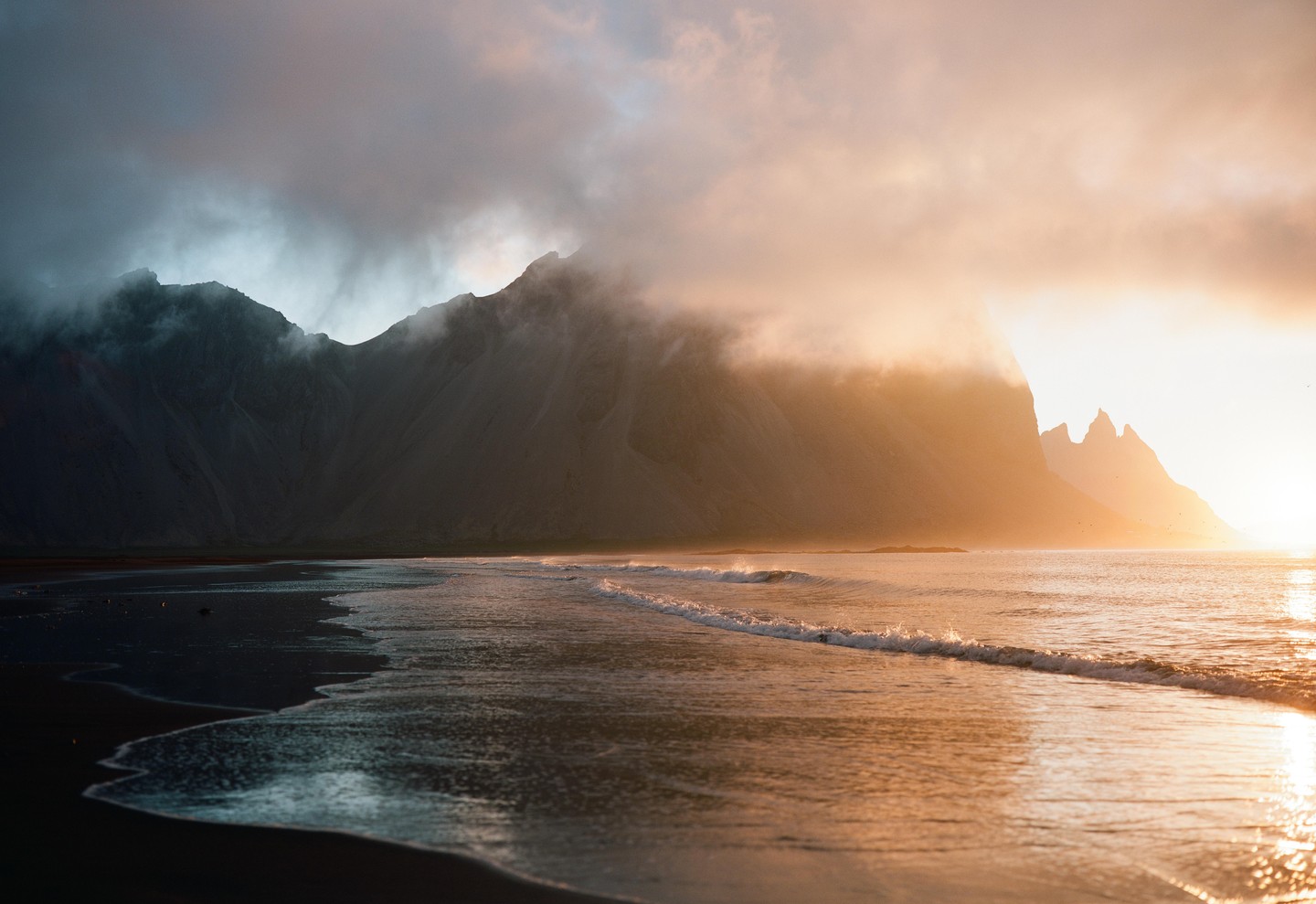 Waves crash on a dark beach with mountains shrouded in fog and golden light in the background.