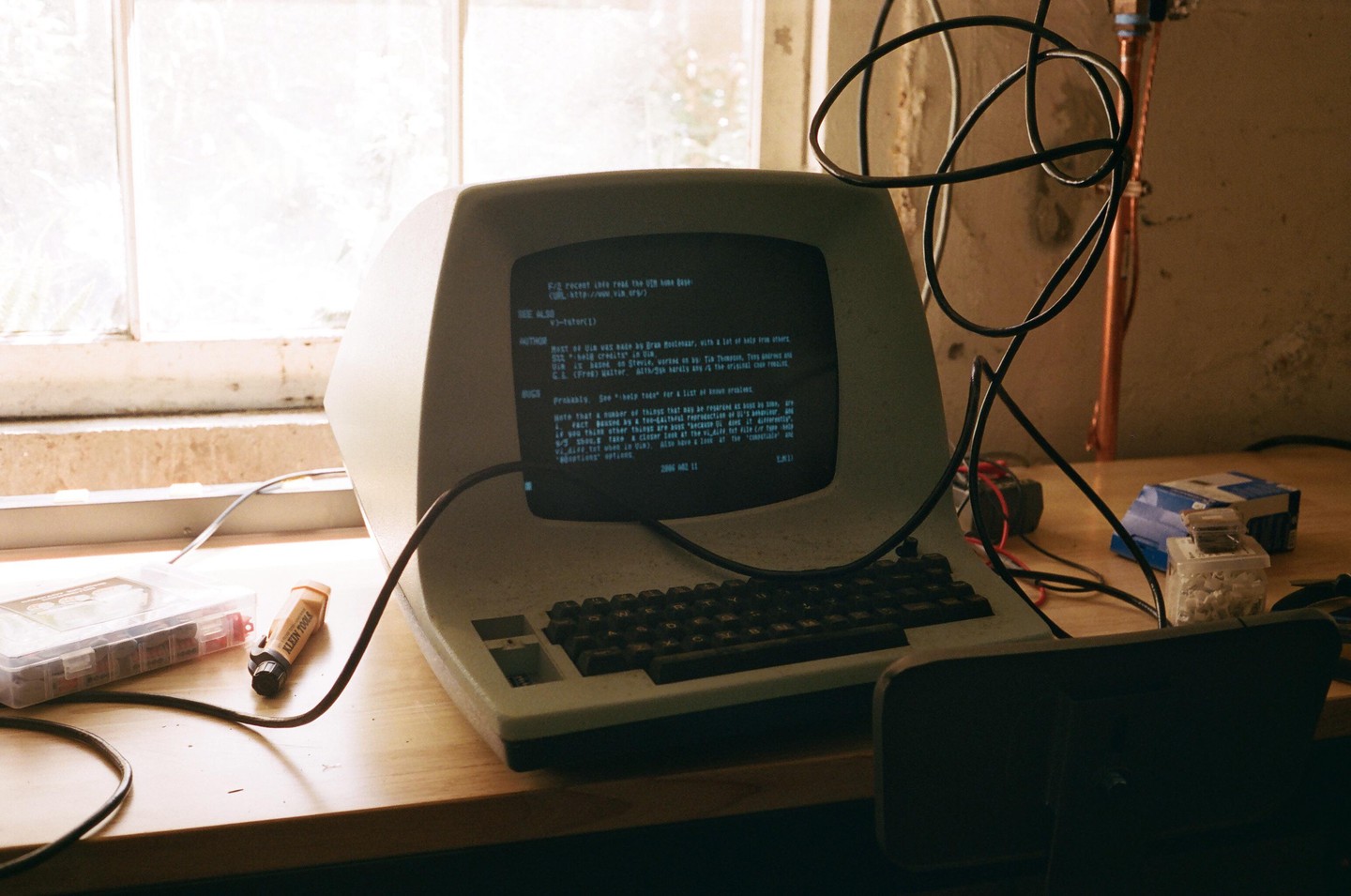 An old computer terminal with a green screen displaying text sits on a wooden desk next to a window.