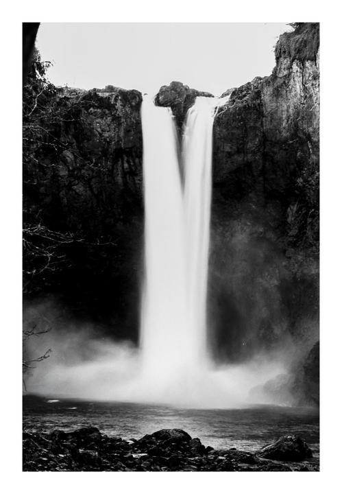 A black and white long exposure shows a waterfall cascading into a misty pool surrounded by dark rocks and cliffs.