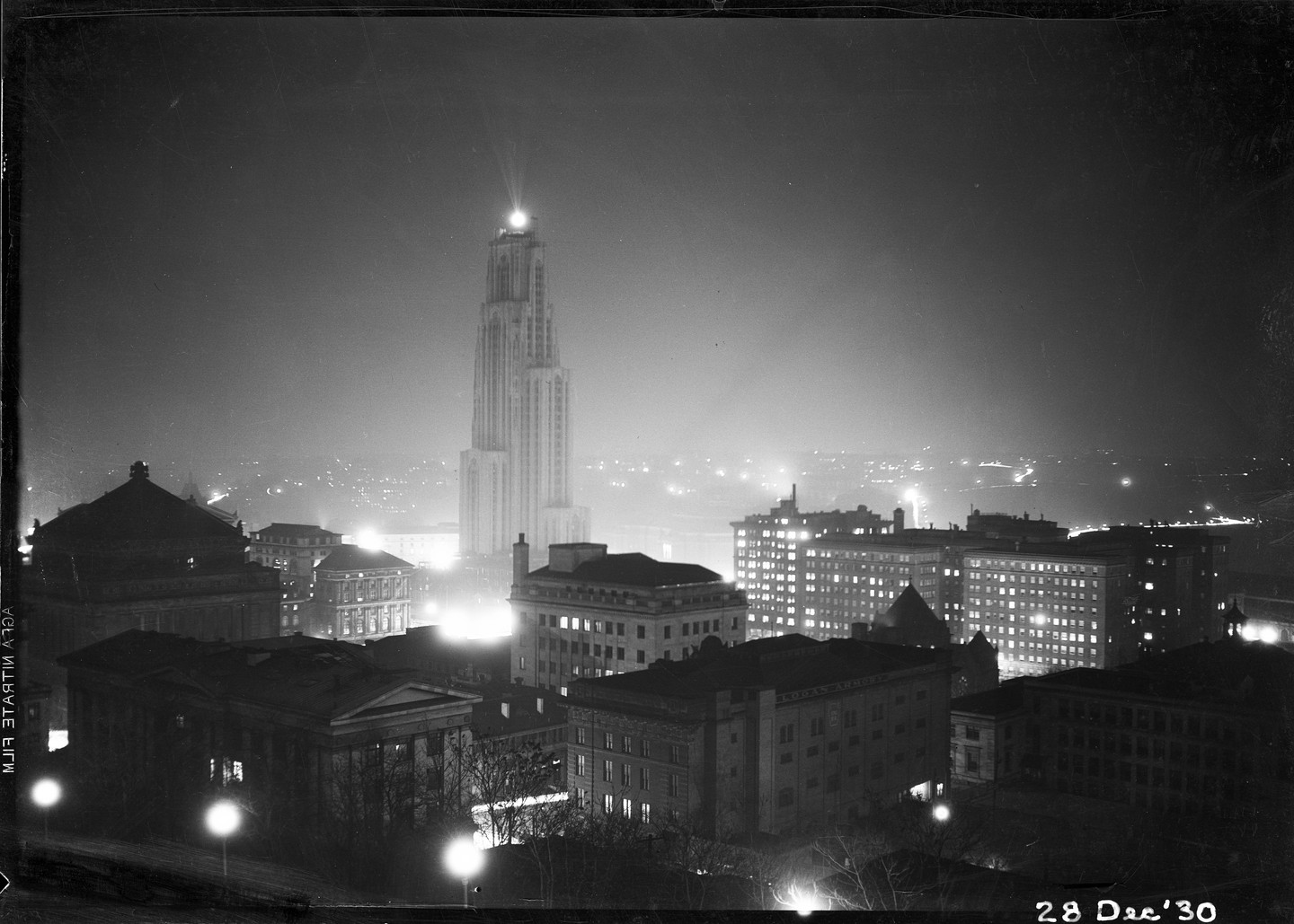 A tall, gothic-style skyscraper with a bright light on top stands prominently among other buildings in a city at night.