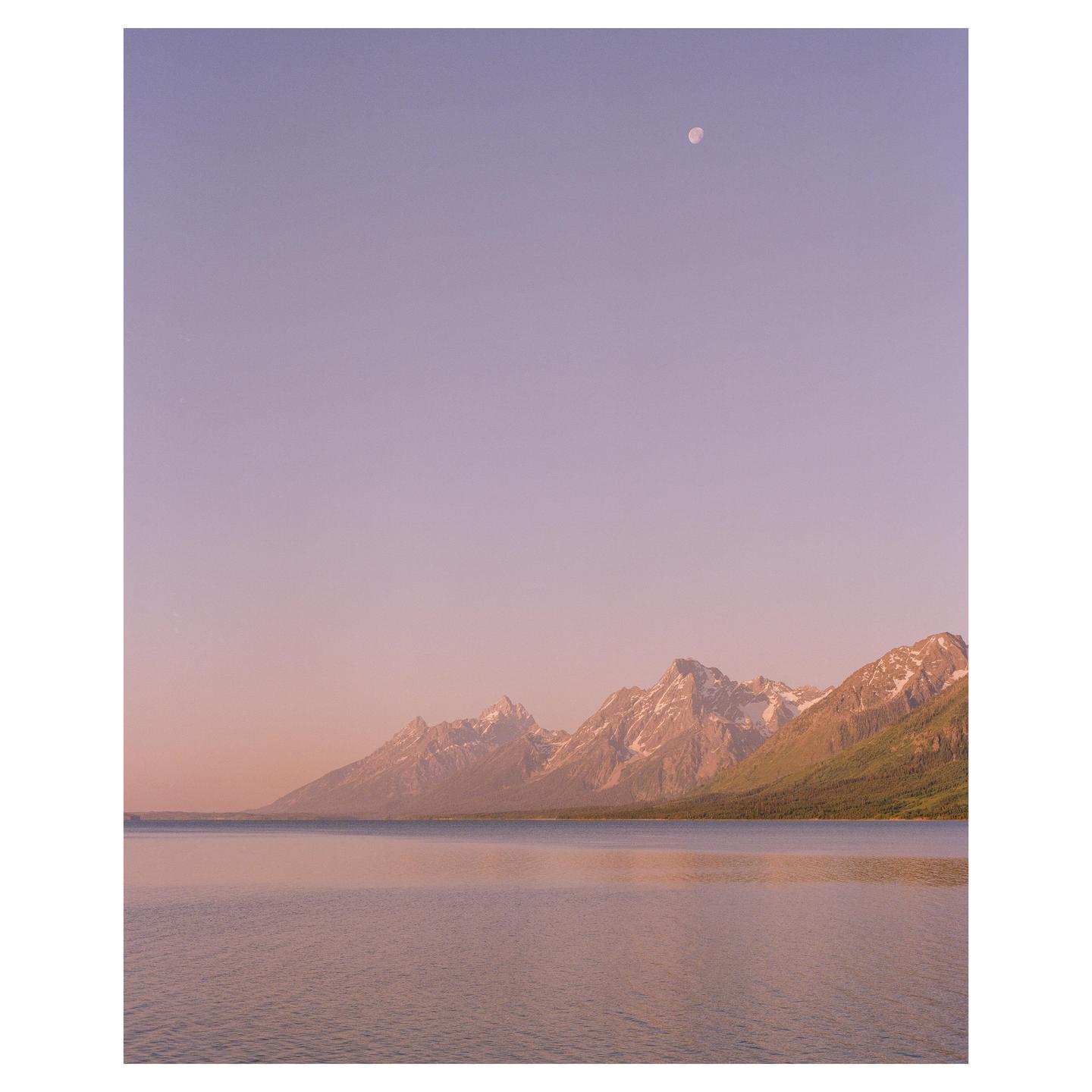 A moon is visible in the sky above a lake and mountains with snow-capped peaks.