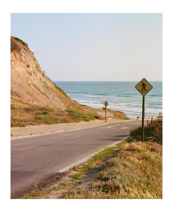 A road descends towards a beach with waves crashing on the shore, next to a cliff and a pedestrian crossing sign.
