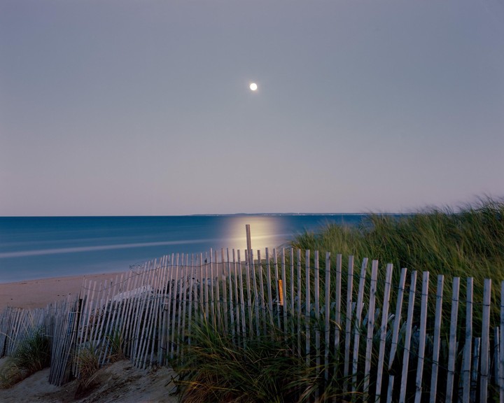 A full moon illuminates the ocean and a wooden fence on a sandy beach at night.