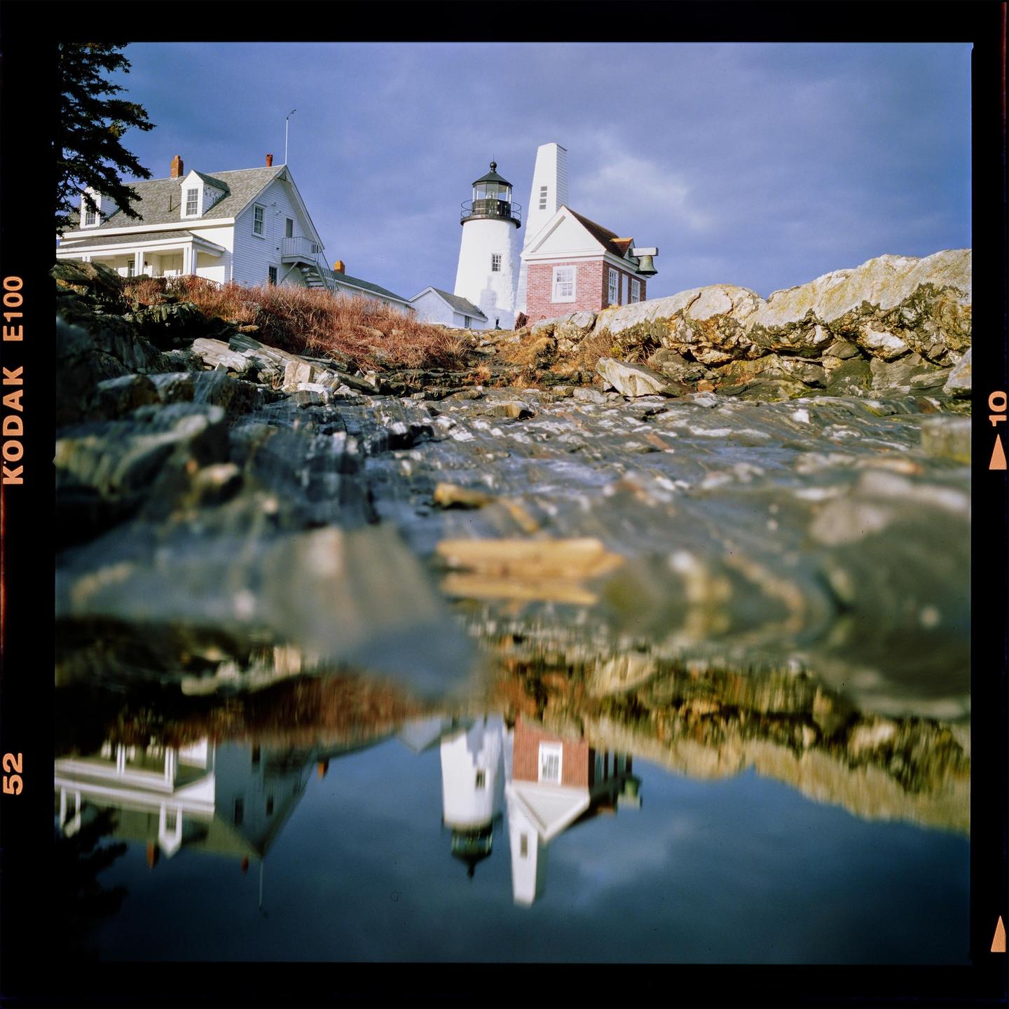 A white lighthouse and house are reflected in a tide pool on a rocky shore under a cloudy sky.