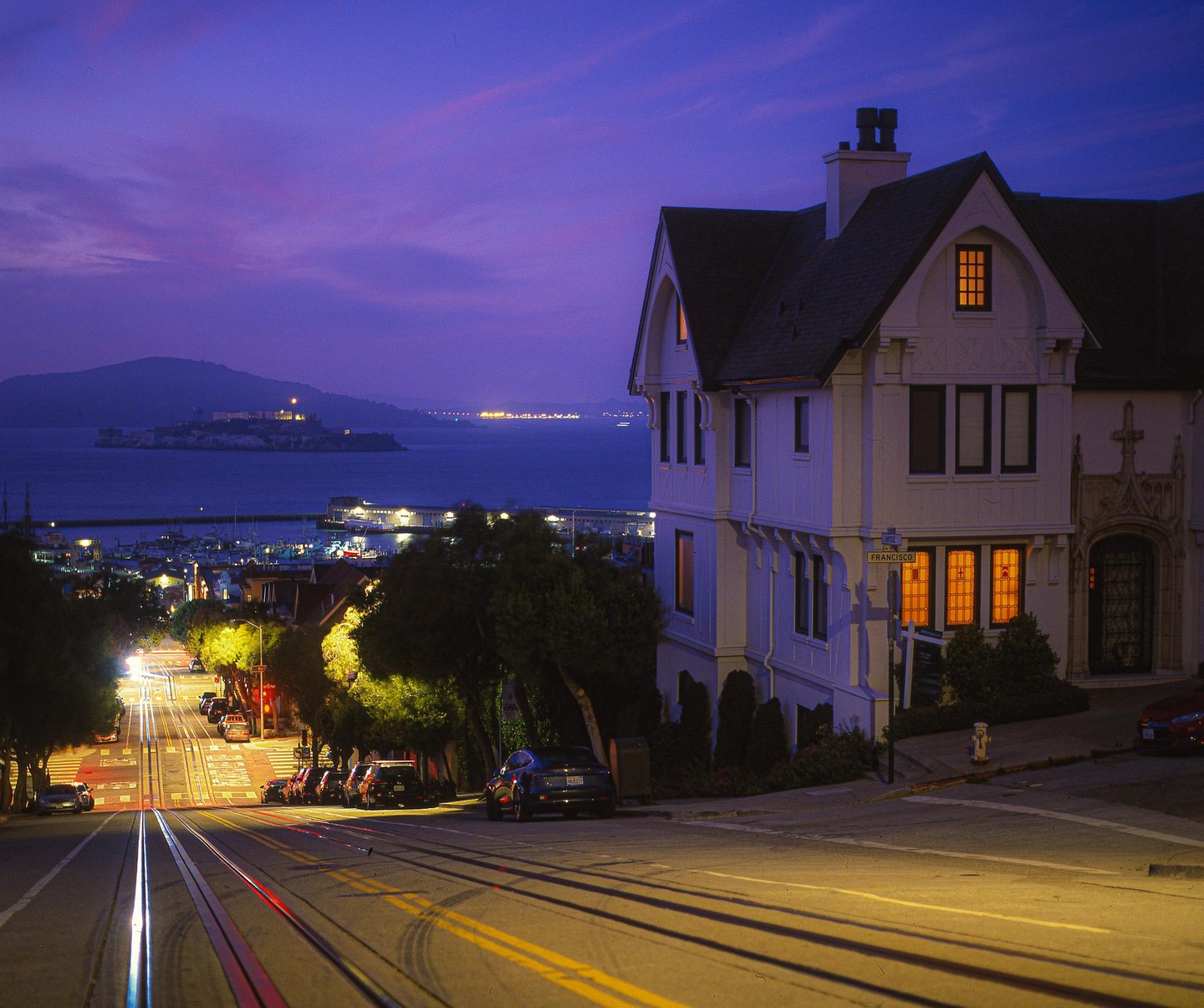A large white house with illuminated windows stands on a hill overlooking a bay with an island and a city street with cable car tracks at dusk.