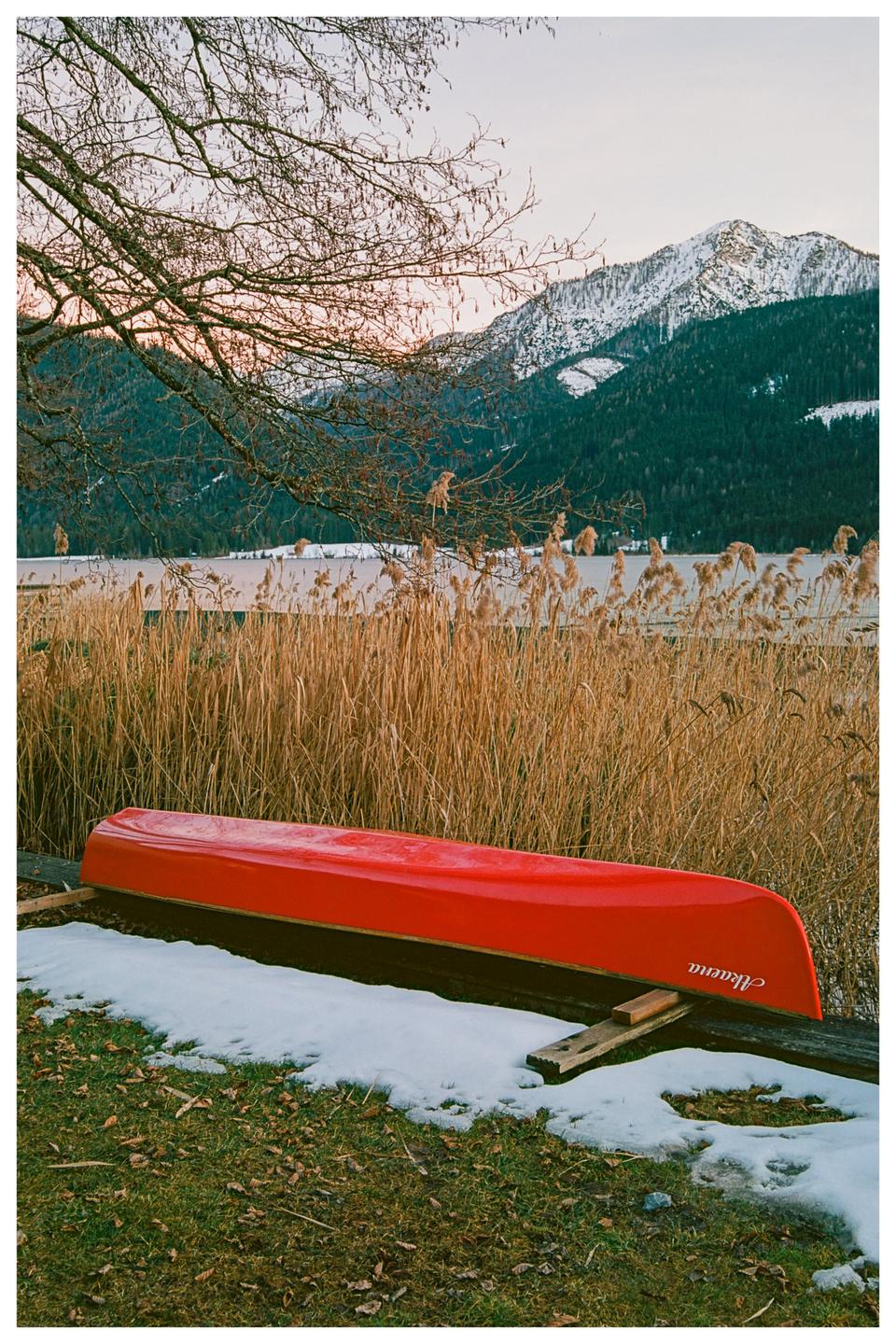 A red canoe rests upside down on a snowy bank beside a lake with tall reeds and snow-capped mountains in the background at sunset.