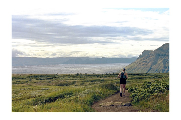 A woman runs on a dirt path through a green field with mountains and a body of water in the background.