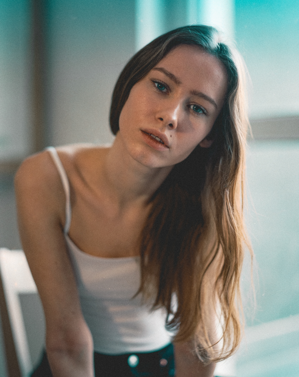 A young woman with long hair and a white tank top looks directly at the viewer.