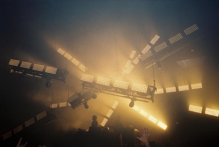A concert stage is illuminated by numerous bright, rectangular stage lights, with a DJ visible in silhouette below.