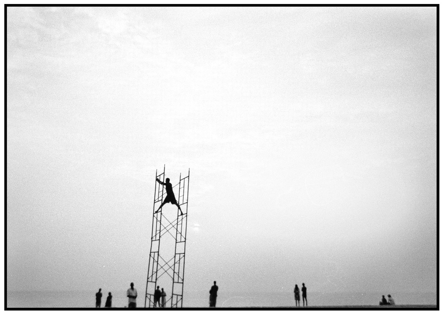A silhouetted man climbs a tall metal structure on a beach with other people in the distance.