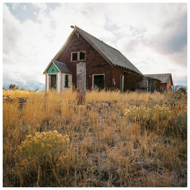 An old, weathered house with a green-painted porch stands in a field of tall, dry grass under a cloudy sky.