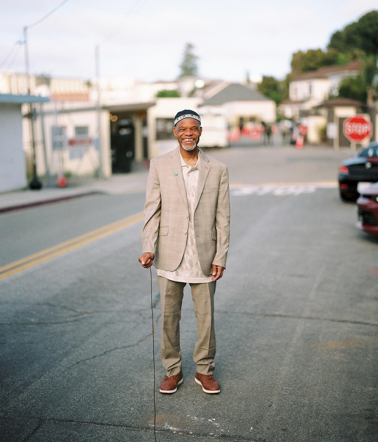 A man in a suit and a patterned cap stands in the middle of a street, holding a thin cord.
