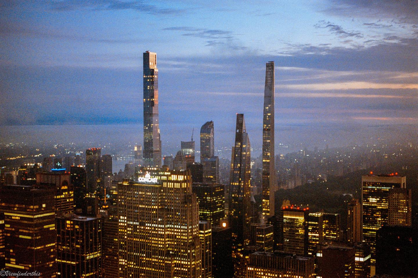 An aerial view of the New York City skyline at dusk shows numerous illuminated skyscrapers and a hazy horizon.
