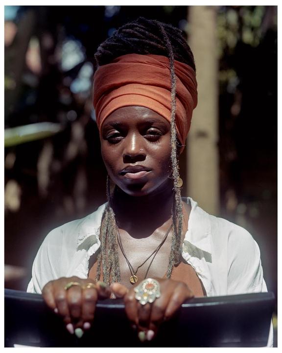 A woman with an orange head wrap and dreadlocks looks directly at the viewer.