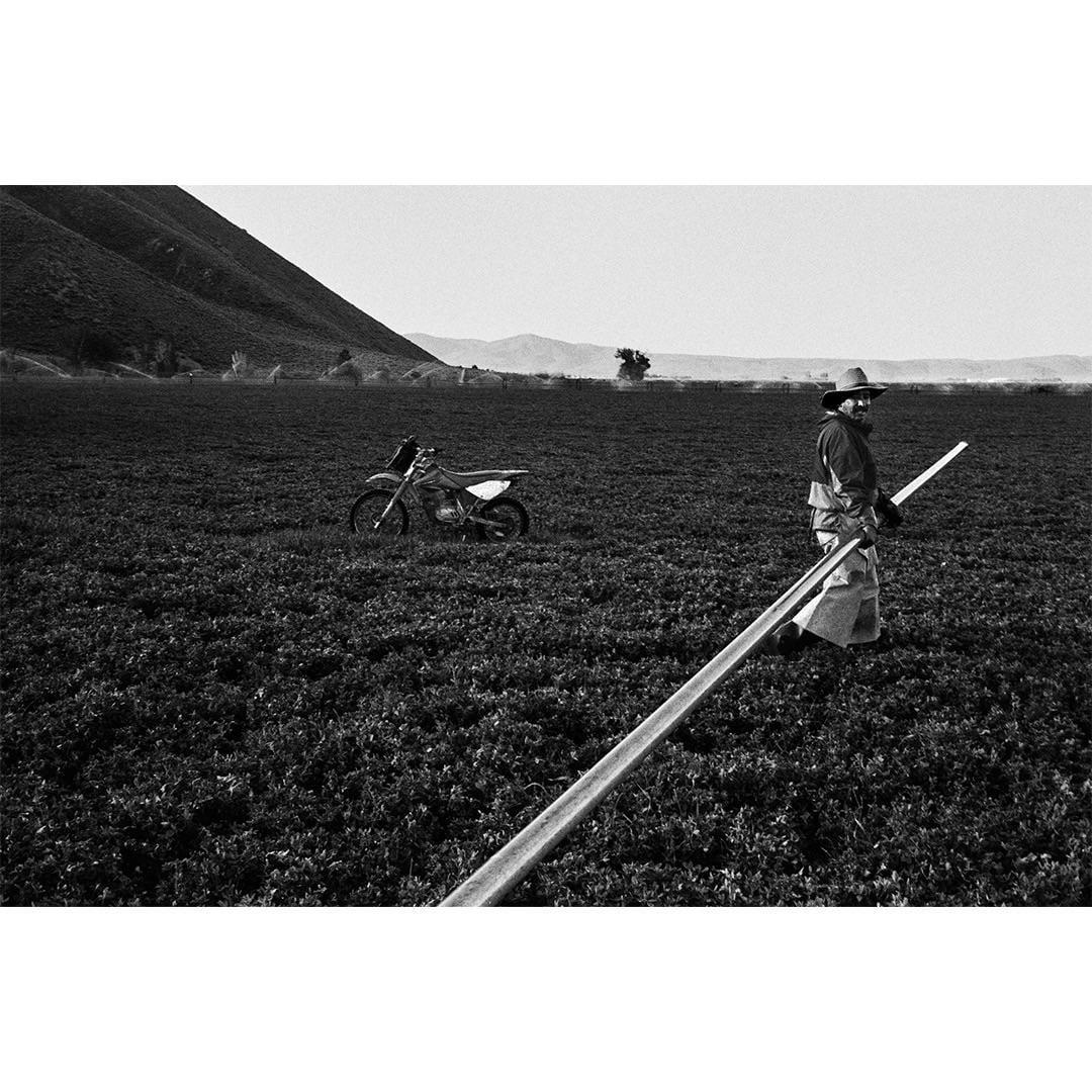 A man in a cowboy hat stands in a field with a motorcycle and a long pipe.