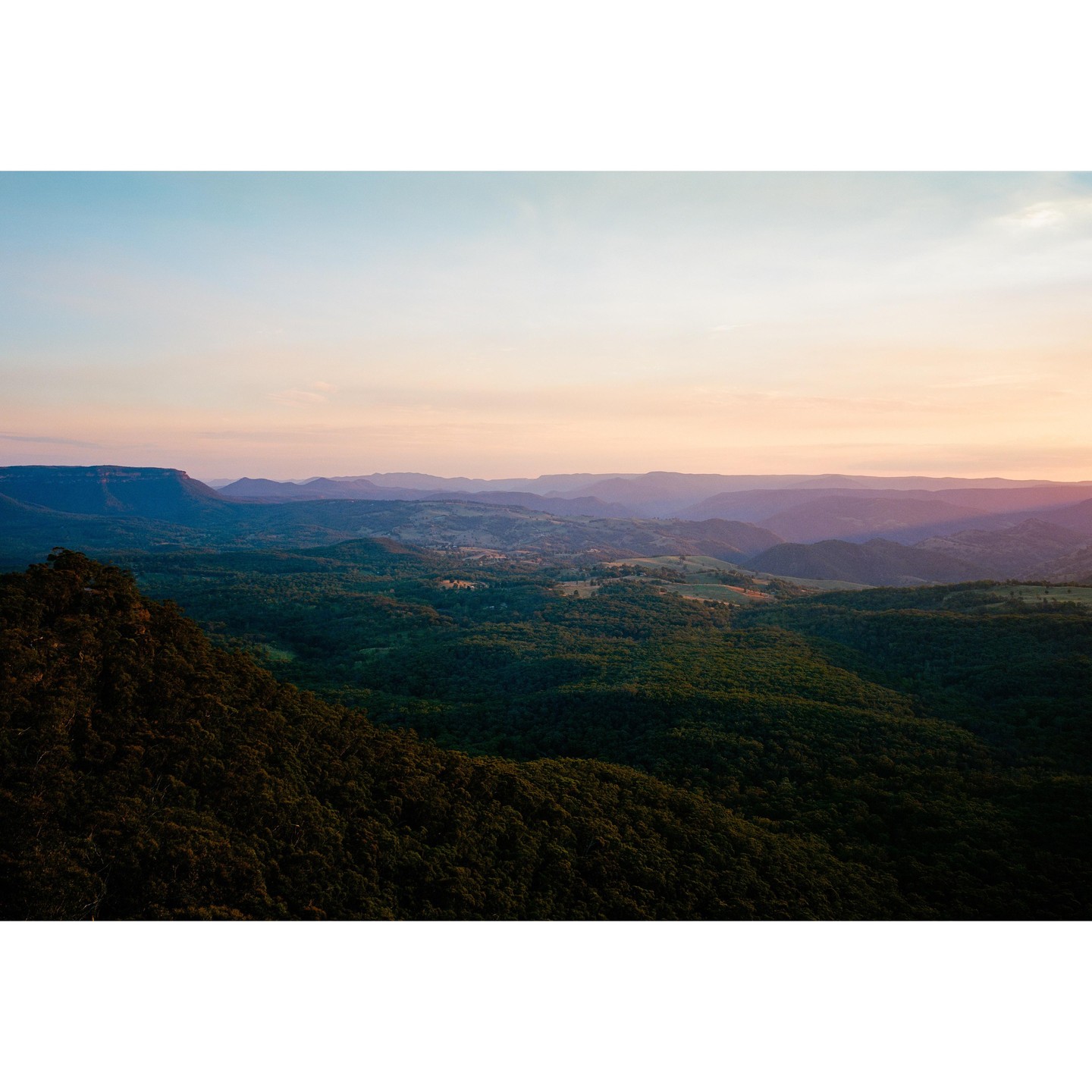 A vast landscape of rolling hills and mountains covered in dense forest under a pastel sky at dusk.