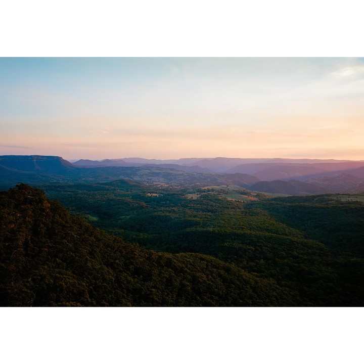 A vast landscape of rolling hills and mountains covered in dense forest under a pastel sky at dusk.