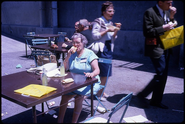 A woman in glasses eats a hot dog at an outdoor table with other people in the background.