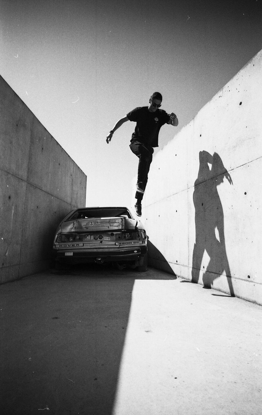 A man jumps over a derelict car between two concrete walls, casting a long shadow on the right wall.