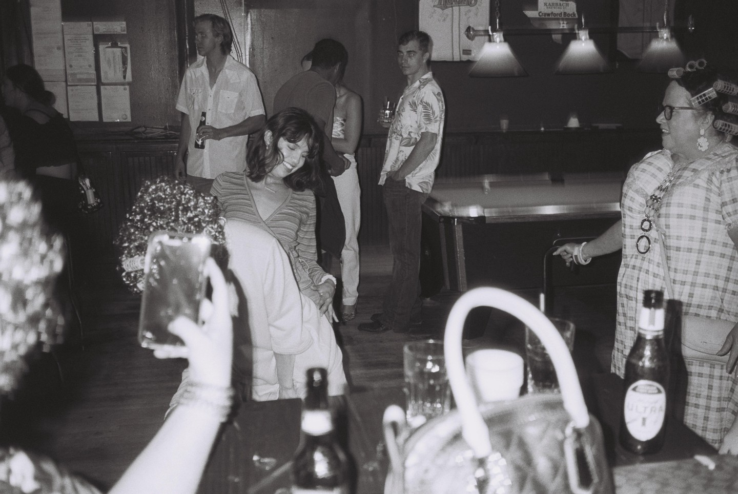 A group of people are gathered in a dimly lit bar with a pool table in the background.