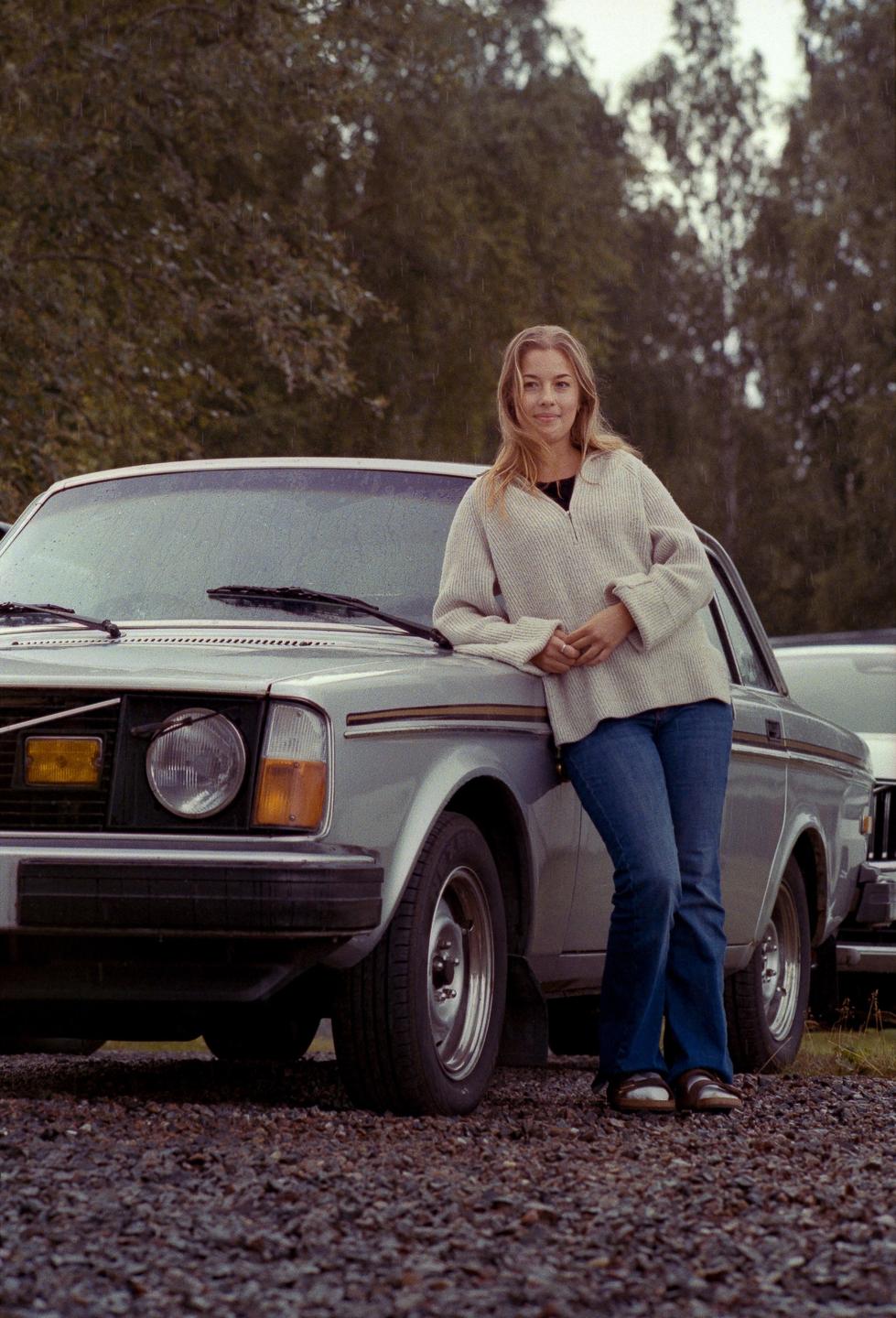 A woman in a sweater and jeans leans against a silver car on a gravel surface with trees in the background.