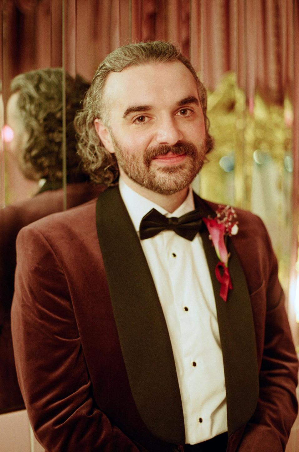 A man in a tuxedo with a boutonniere smiles at the camera, with his reflection visible in a mirror behind him.