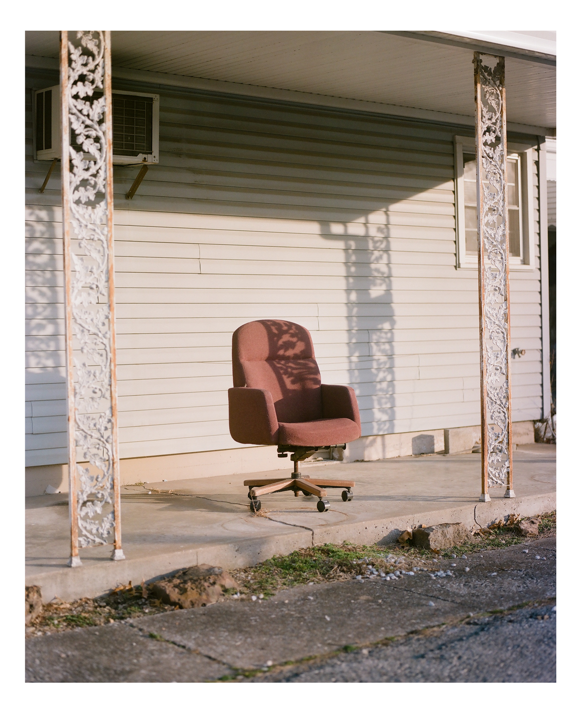 An office chair sits on a porch with decorative pillars and an air conditioning unit.