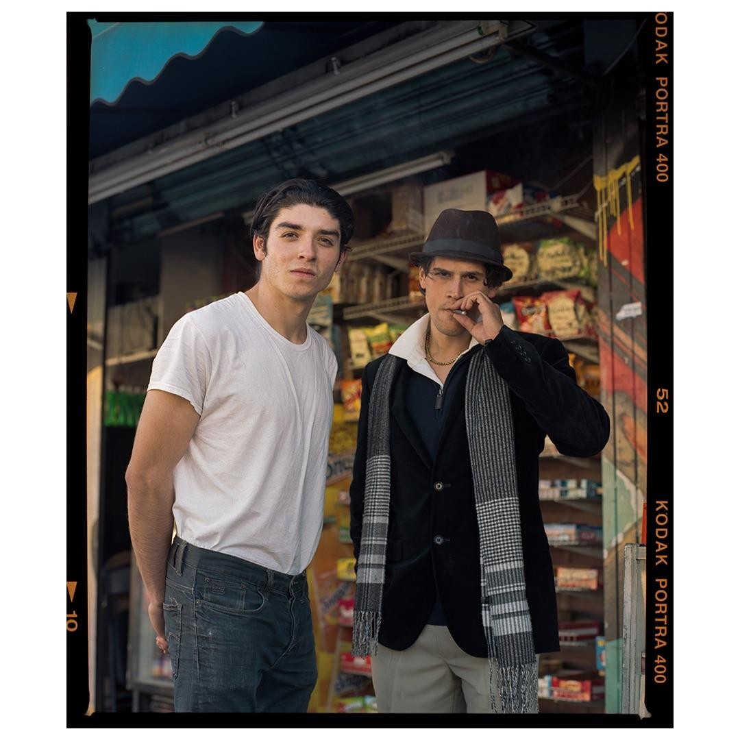 Two men stand in front of a store, one smoking a cigarette.