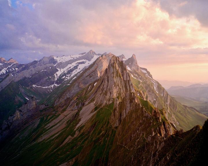 A mountain range with snow-capped peaks and green slopes is illuminated by the golden hour light under a cloudy sky.