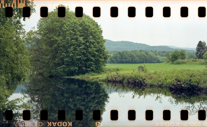 A river reflects trees and a small shed in a green landscape under a bright sky.