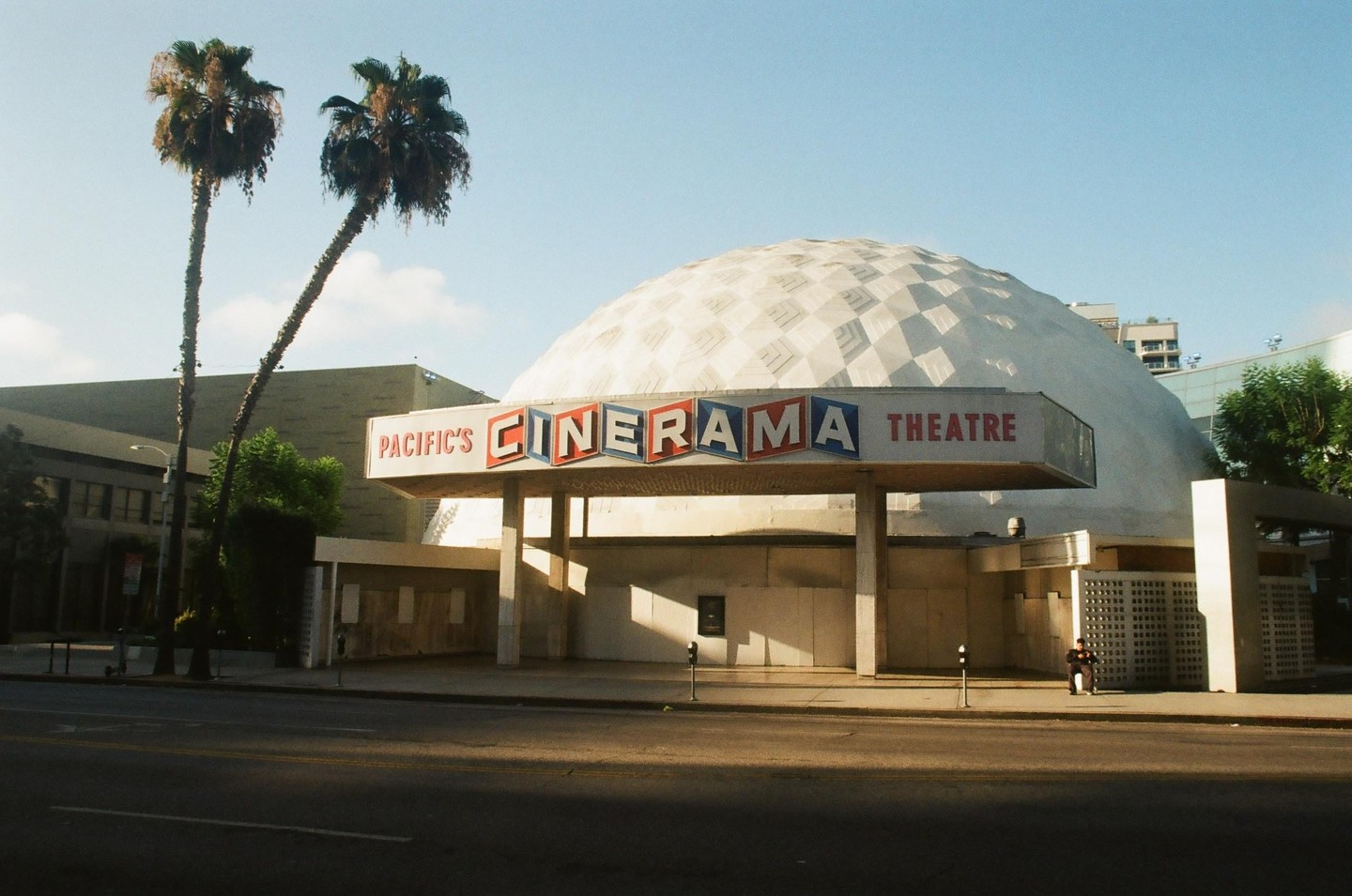 A large domed building with a sign that reads "Pacific's Cinerama Theatre" stands next to a street with two palm trees.