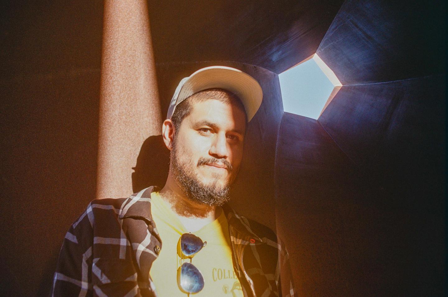 A man wearing a cap and sunglasses on his shirt is illuminated by a shaft of light in a dark, angular space.