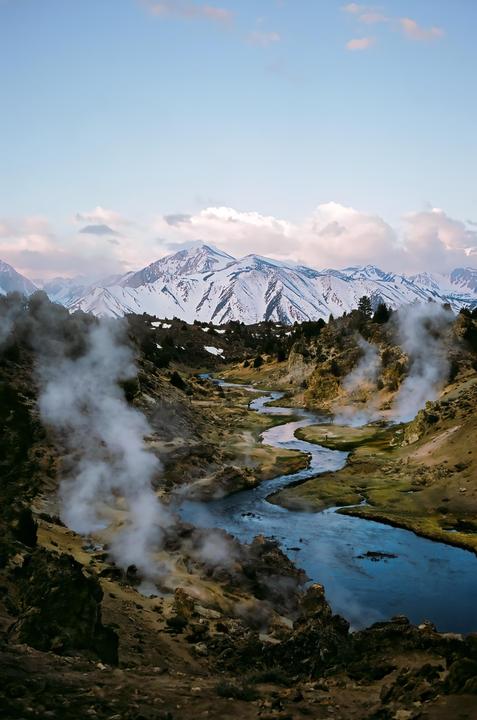 A winding river flows through a valley with steam rising from the ground and snow-capped mountains in the distance under a cloudy sky.