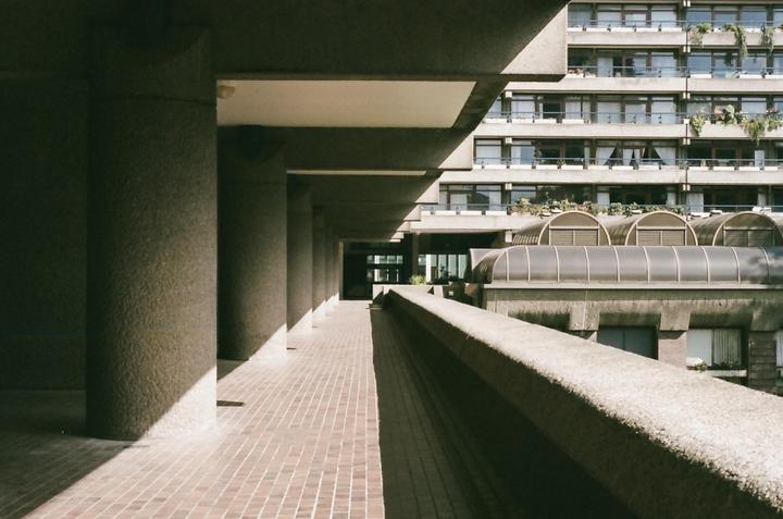 A long concrete walkway with pillars on the left and a concrete railing on the right leads to a building with many windows and balconies.
