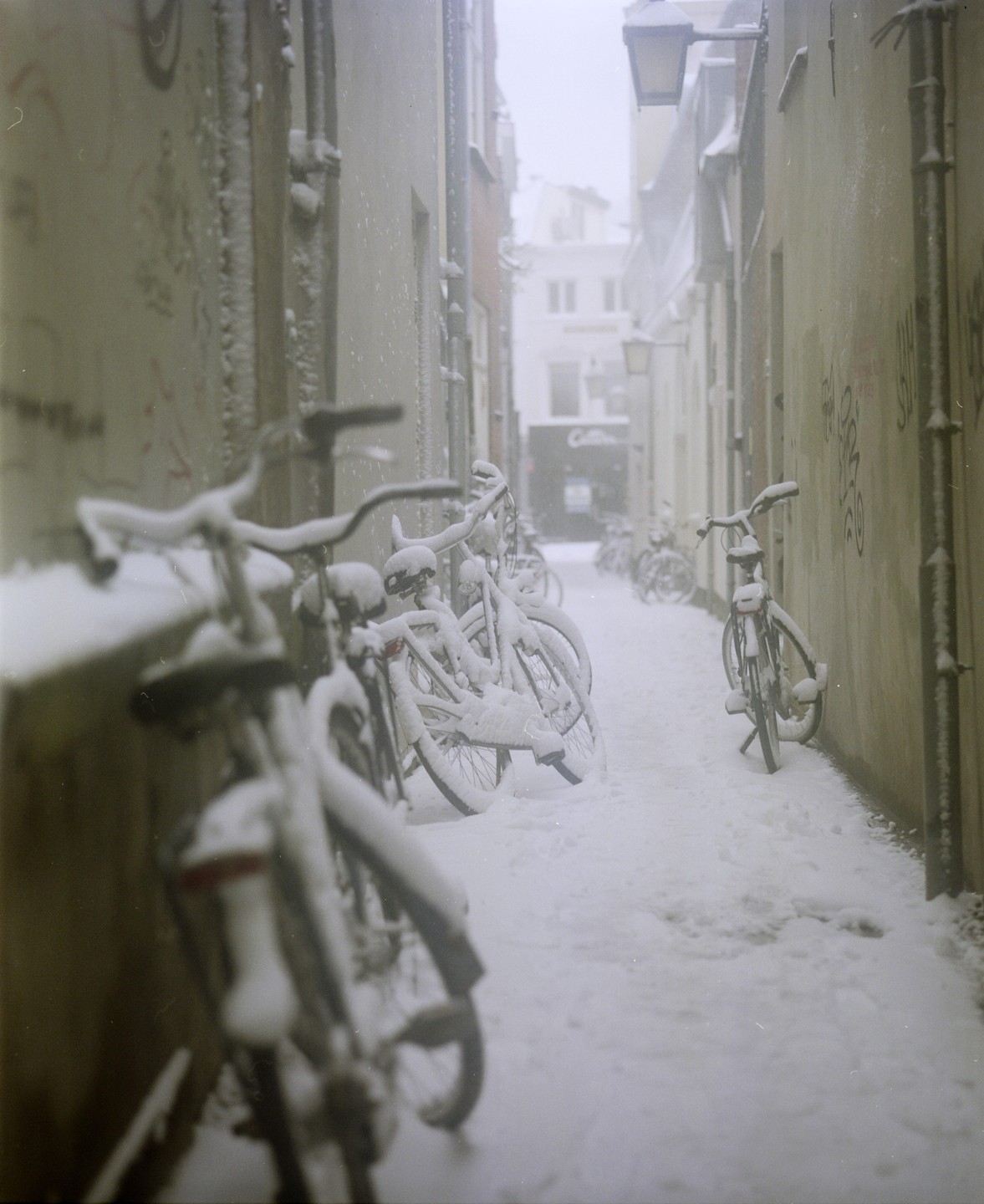 Bicycles covered in snow line a narrow alleyway in a city.
