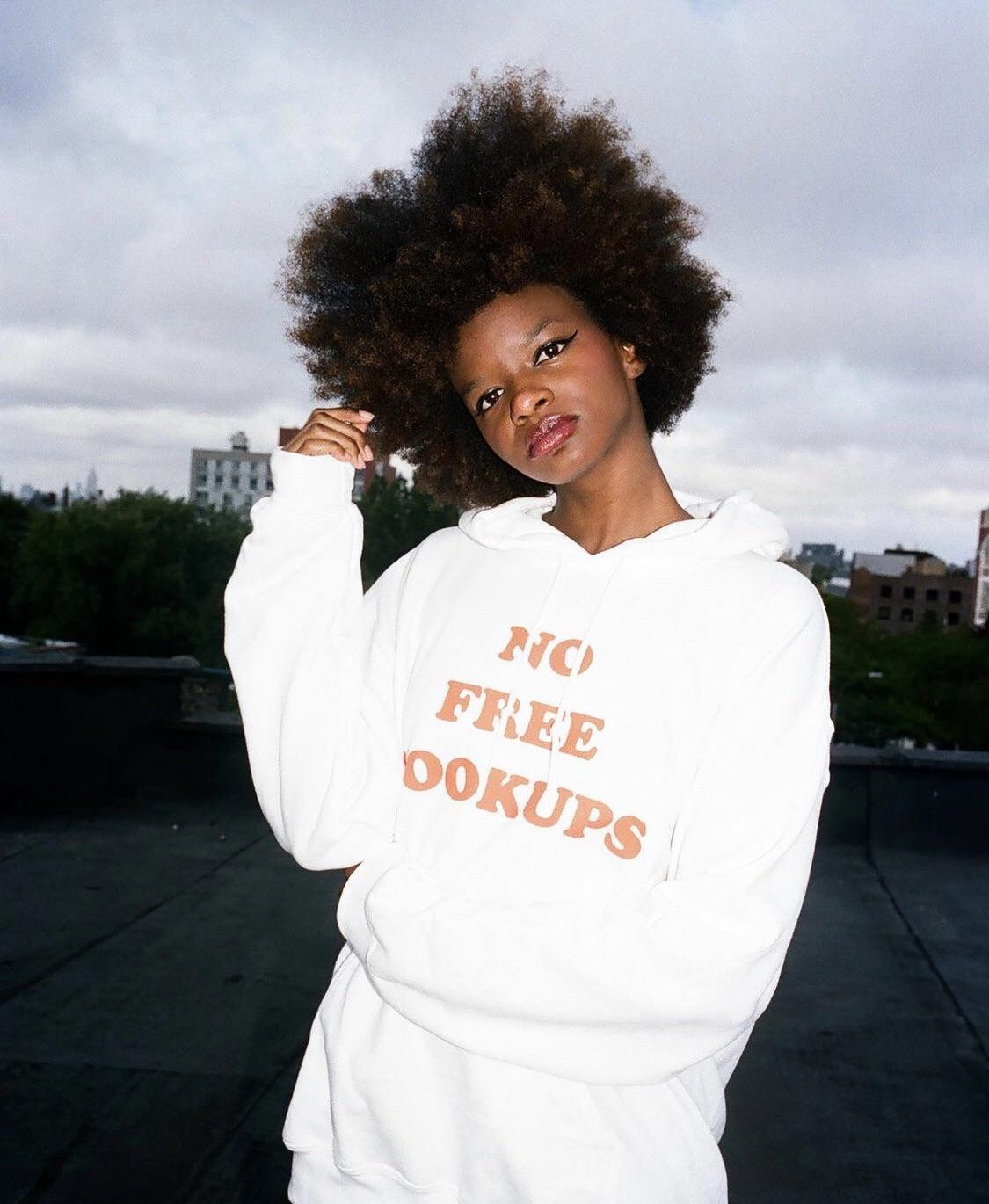 A woman with an afro wears a white hoodie with orange text on a rooftop.