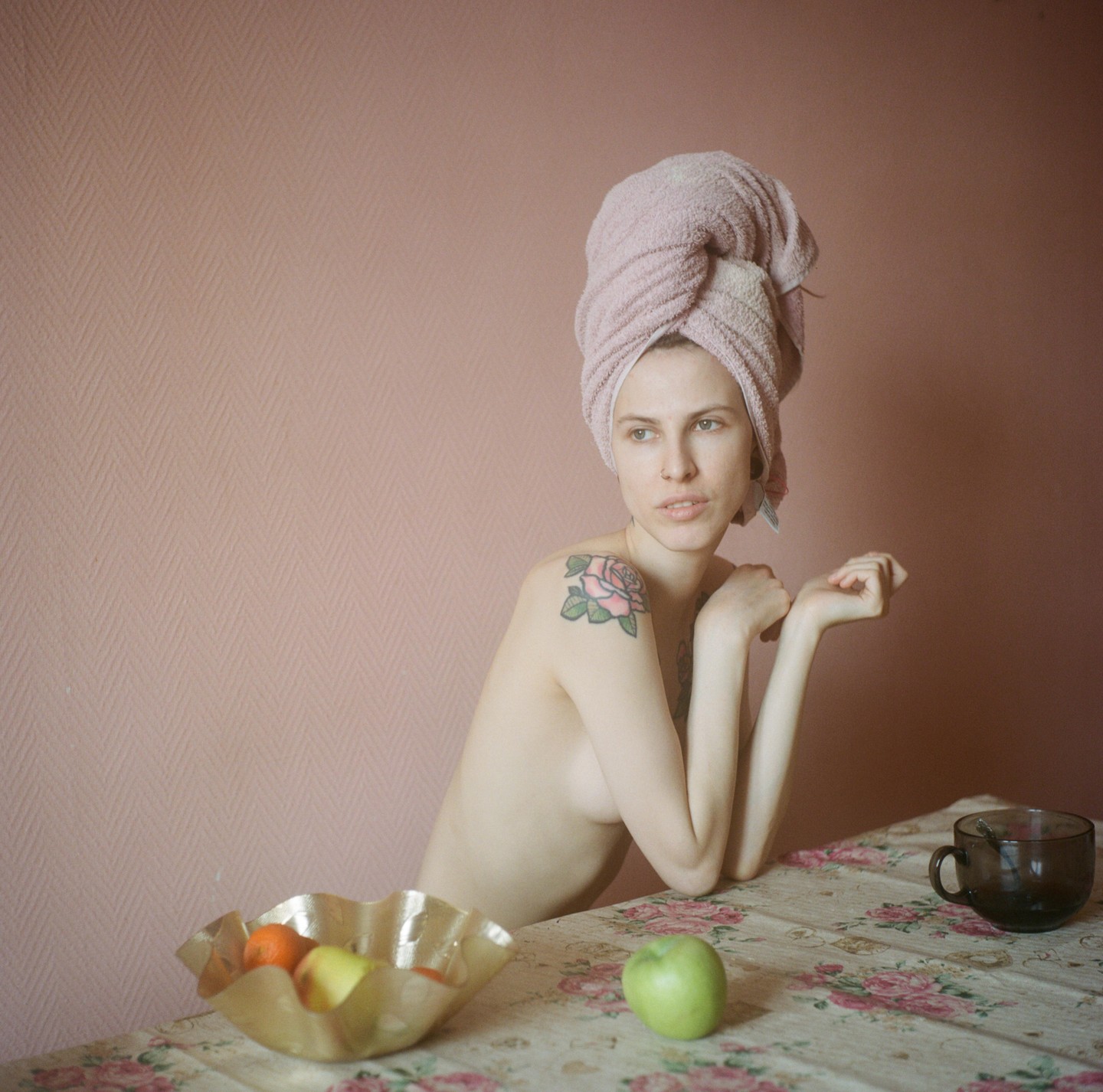 A woman with a towel wrapped around her head sits at a table with a bowl of fruit and a cup.