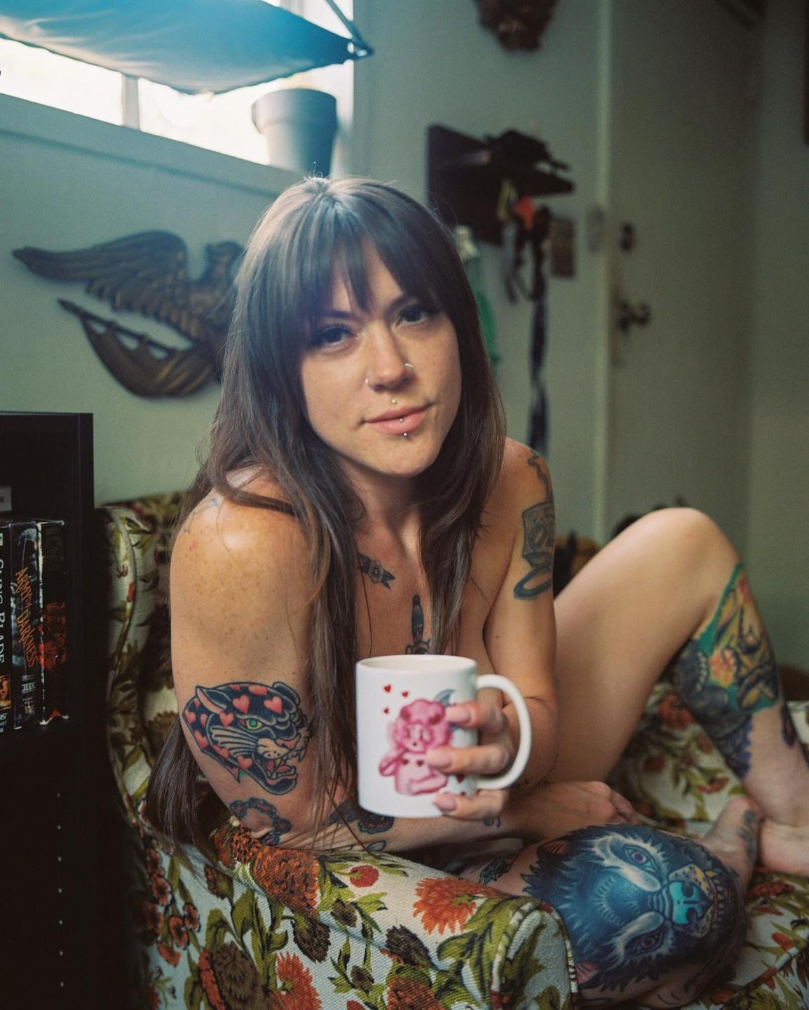 A woman with tattoos and piercings sits in a floral armchair, holding a mug.