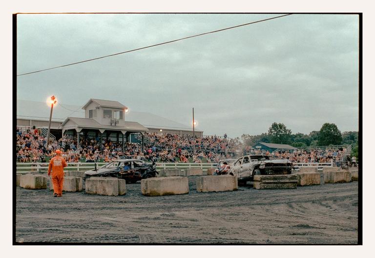 A man in an orange jumpsuit walks past wrecked cars in an arena with a crowd watching from bleachers.
