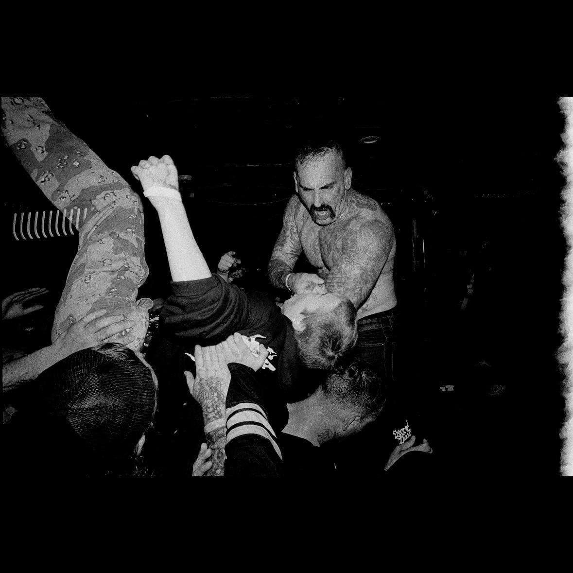 A man with a mustache and tattoos is screaming while holding another man by the head in a dark, crowded room.