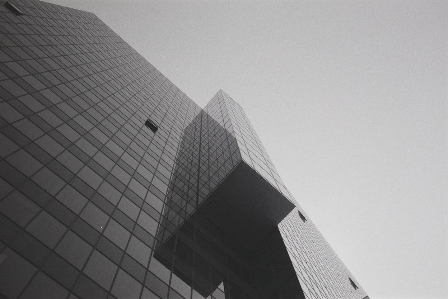 A low-angle view shows the glass facade of a modern building reflecting another building against a bright sky.