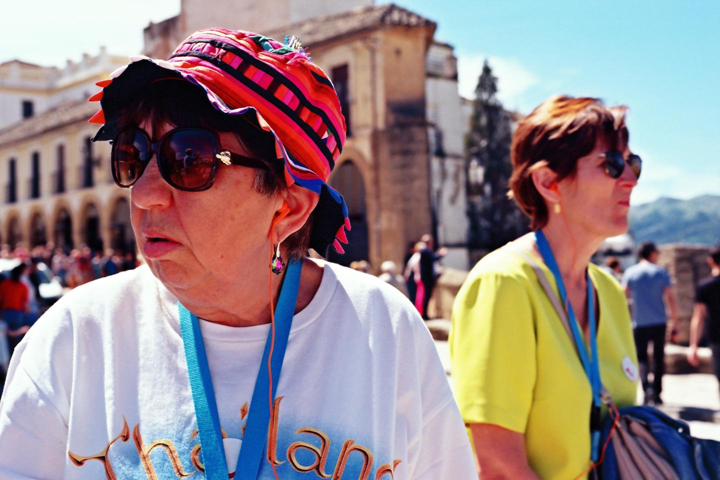 Two women wearing sunglasses and lanyards stand outdoors with historic buildings in the background.