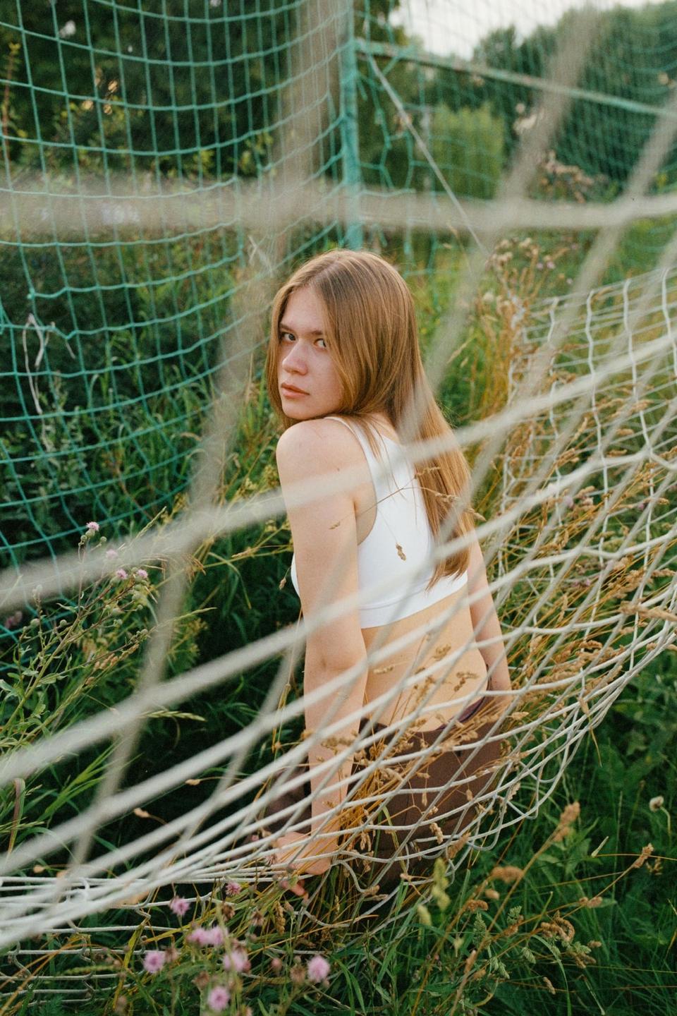 A woman with long hair looks over her shoulder through a soccer net in a field of tall grass.