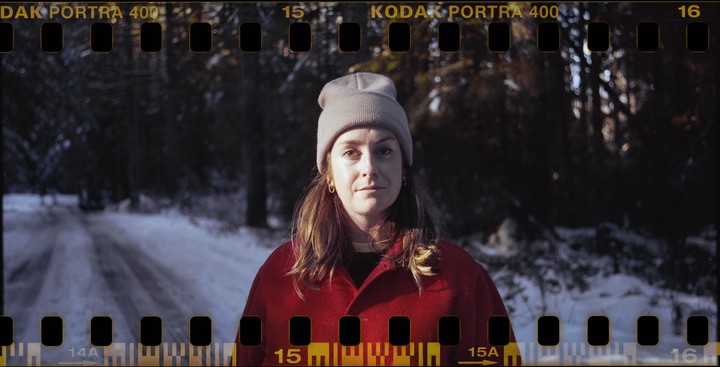 A woman in a red coat and beanie stands on a snowy road in a forest.