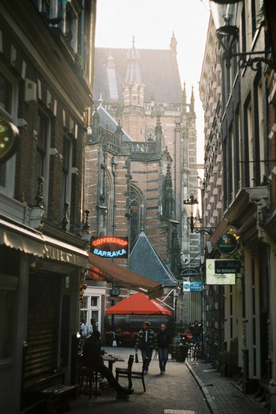 A narrow street in Amsterdam is lined with shops and restaurants, with a large historic building visible in the background.