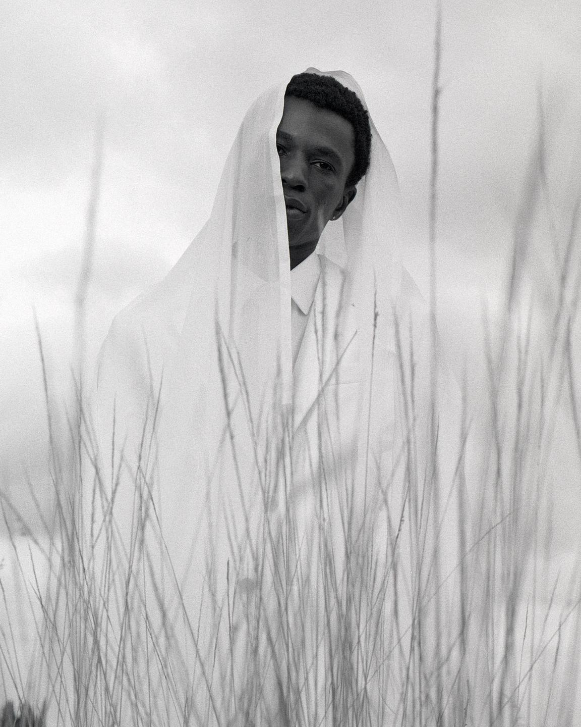 A man with a white veil over his head stands in a field of tall grass.