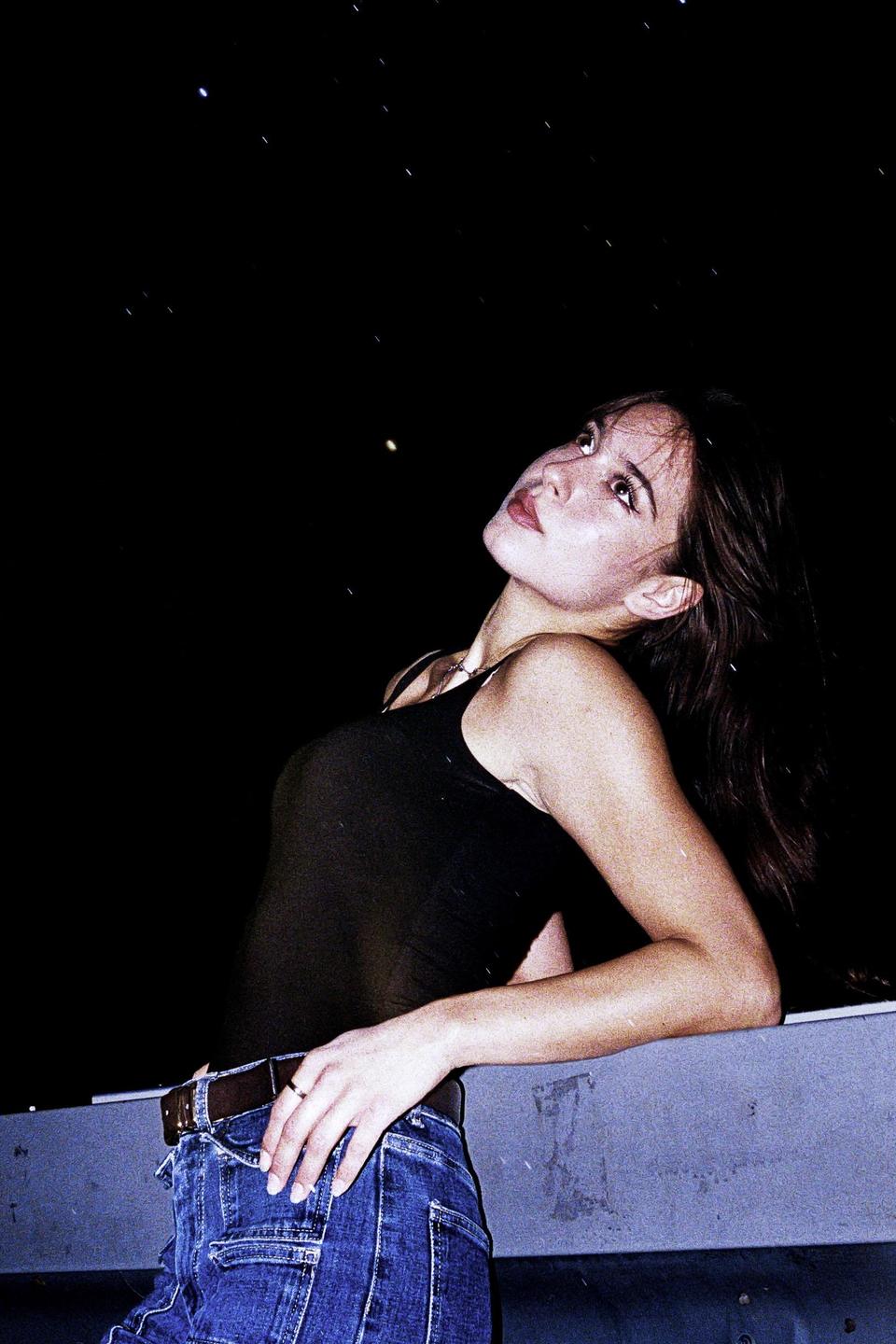 A woman in a black tank top and jeans looks up at a starry night sky.