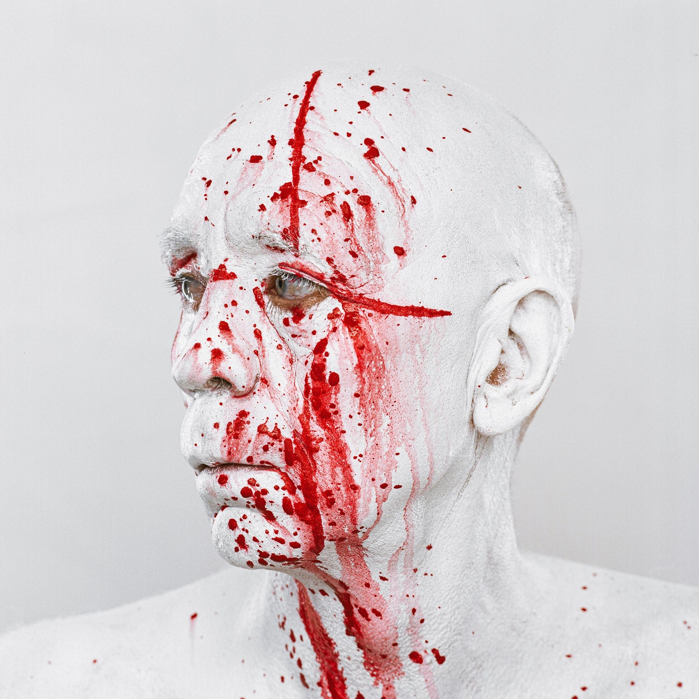 A person with white body paint and red splatters on their face and shoulder looks to the left.