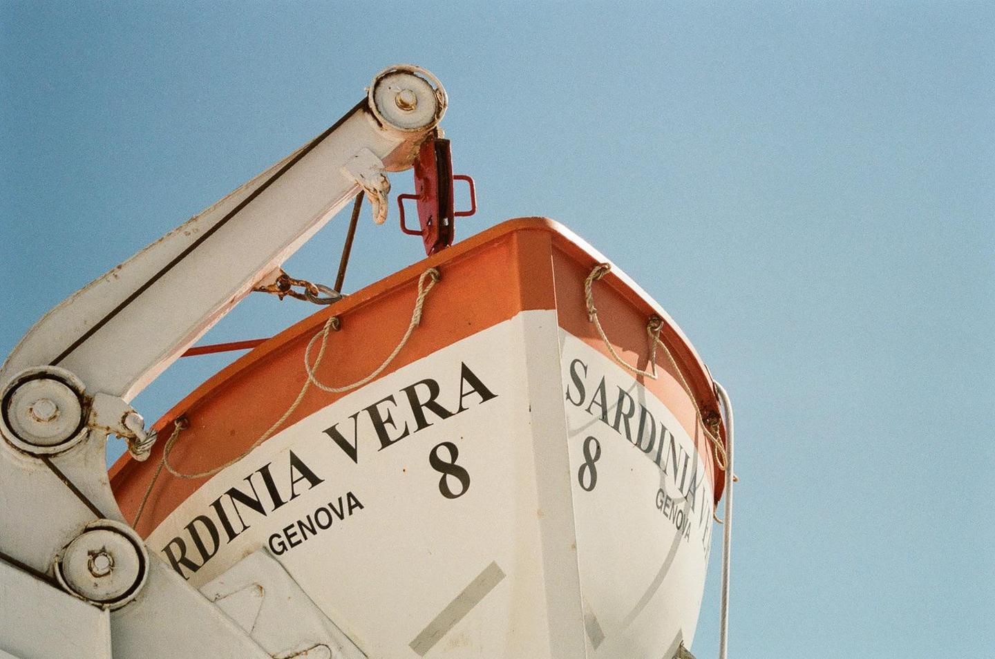 A white and orange lifeboat with "SARDINIA VERA" and "GENOVA" written on its side is suspended against a clear blue sky.